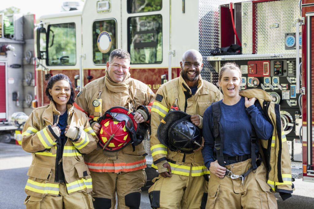 A multi-ethnic group of four firefighters standing together in front of fire engines, wearing fire protection suits and holding helmets. Two of them are women. They are looking at the camera, smiling.